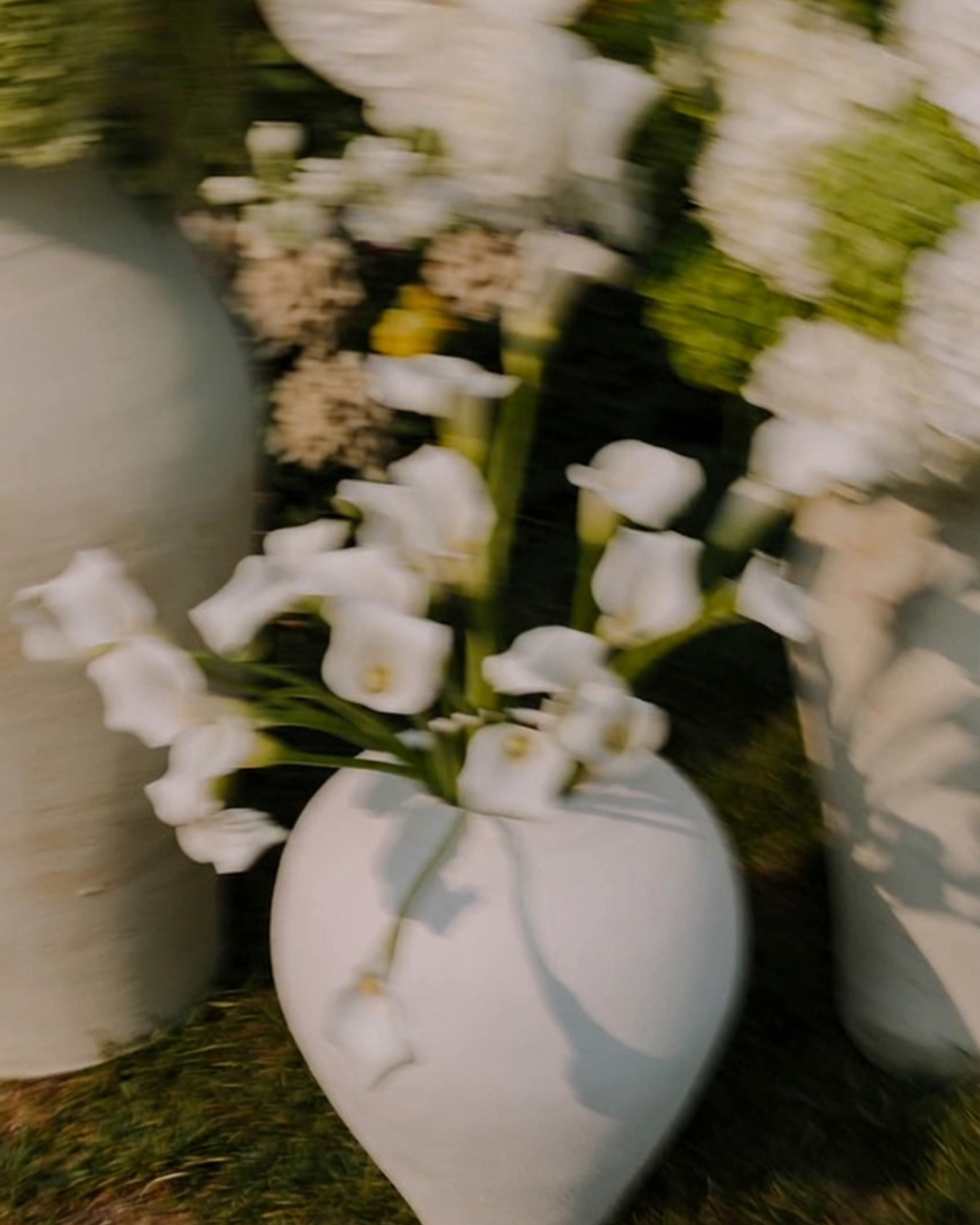 White ceramic heart-shaped vase filled with white calla lilies, photographed outdoors on grass with blurred floral arrangements in the background.
