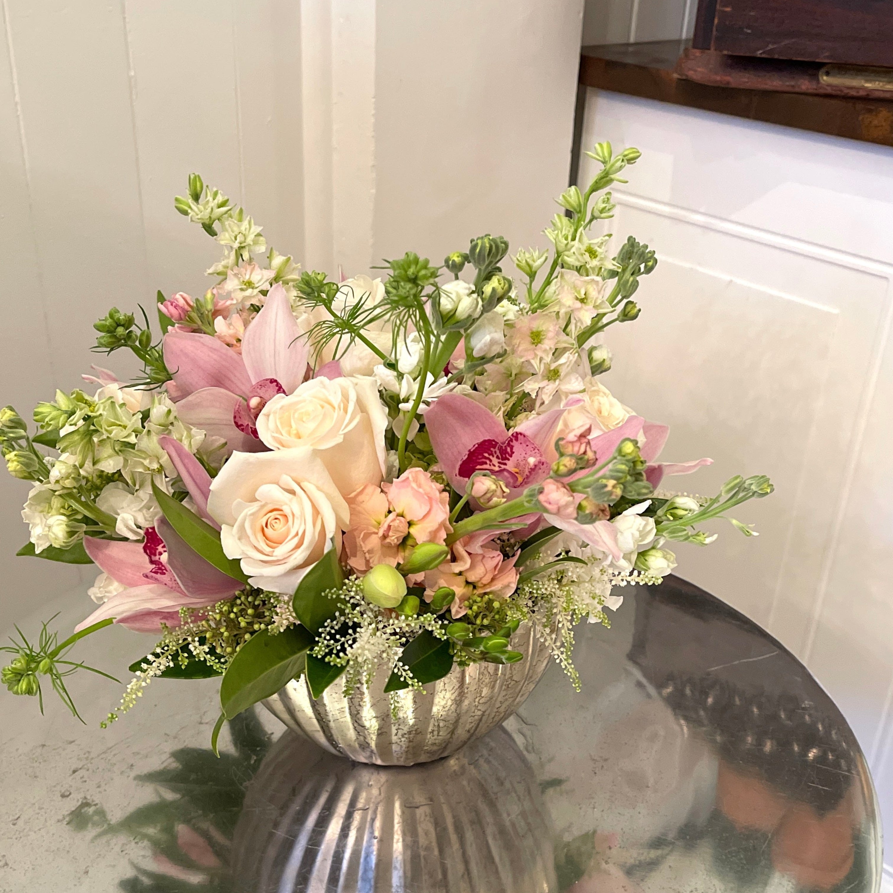 Soft pastel floral arrangement in a silver vase on a reflective surface with a neutral background