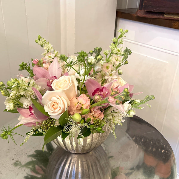 Soft pastel floral arrangement in a silver vase on a reflective surface with a neutral background