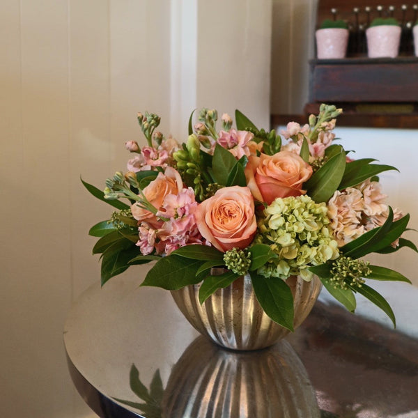 Bouquet of bright/coral flowers in a metallic vase on a reflective surface with a neutral background